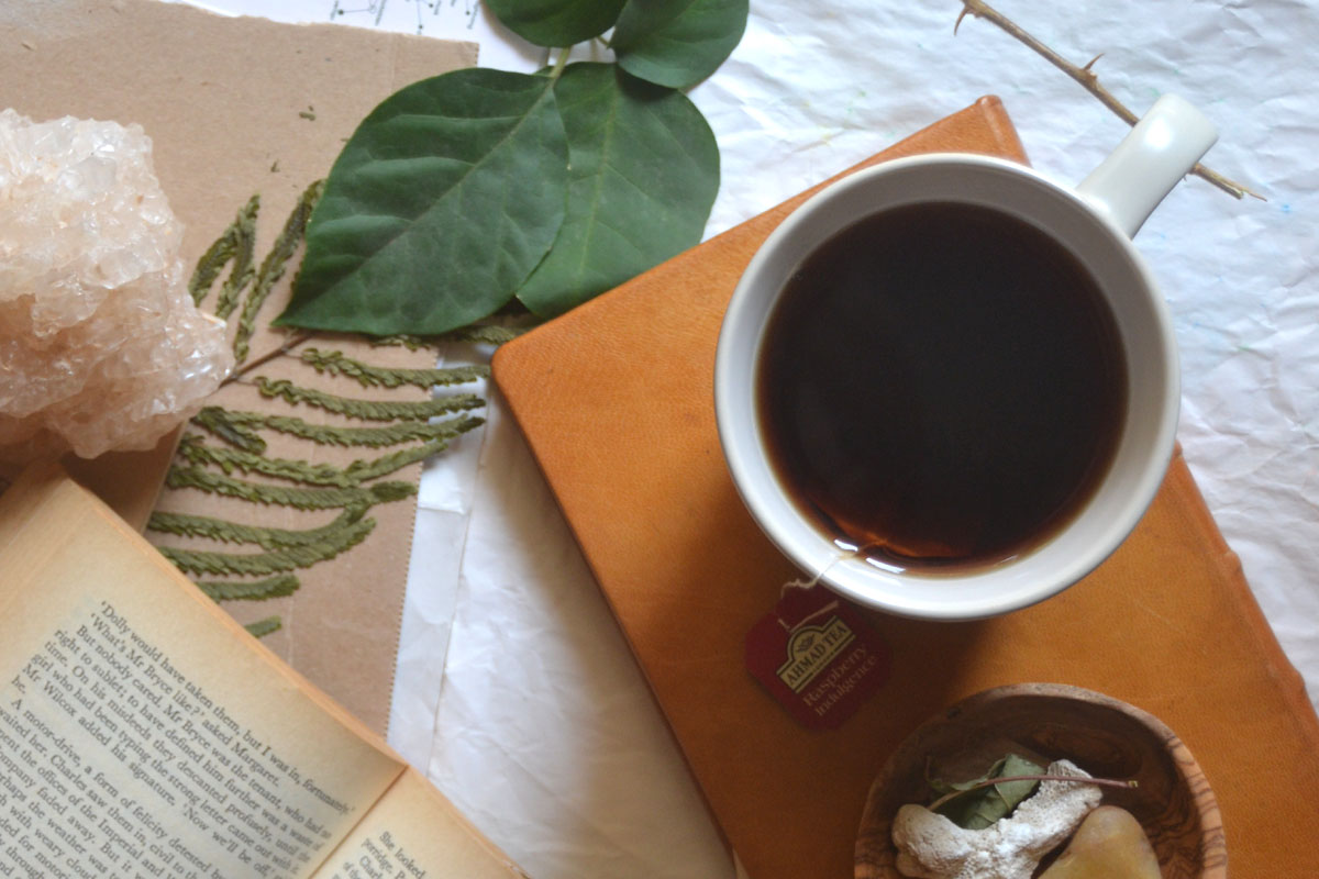 A ceramic mug of herbal tea resting on a closed paper notebook next to an open book to illustrate the simple pleasures of a slow lifestyle.