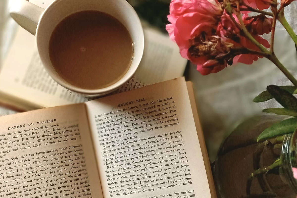 An open novel resting on a rustic wooden table next to a warm cup of coffee and fresh wildflowers in a vase for a slow-paced morning.