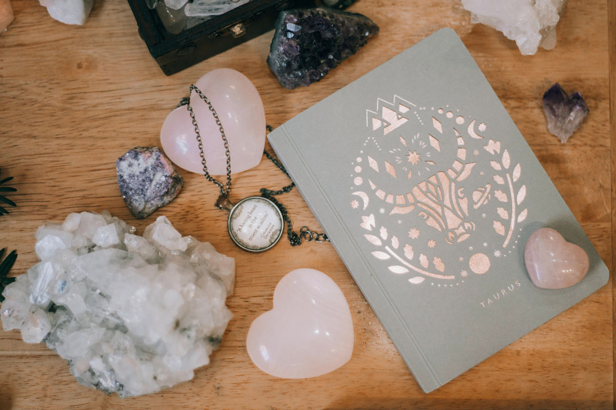 A grey journal lays on a wooden desk alongside pink and white crystals.