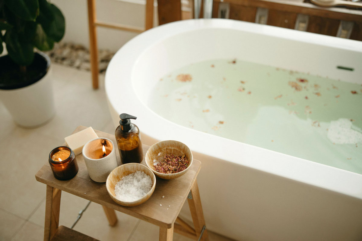 A cozy bathtub surrounded by lit candles, dried rose petals, and botanical element details for a relaxing self-care ritual.