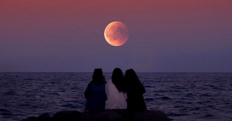 Three female friends sitting on coastal rocks watching a giant red full moon rise over the ocean at sunset, practicing a mindful full moon ritual.