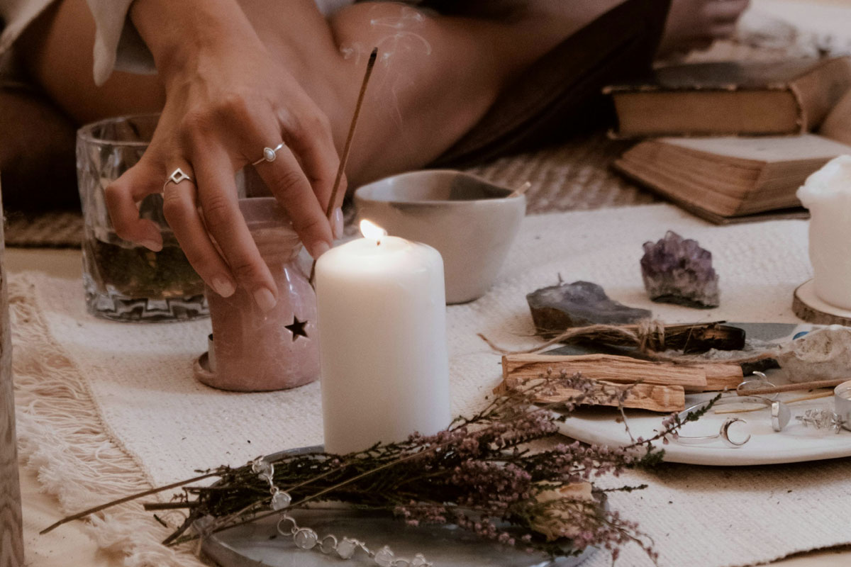 A woman sits on the ground surrounded by herbs and lights a stick of incense.