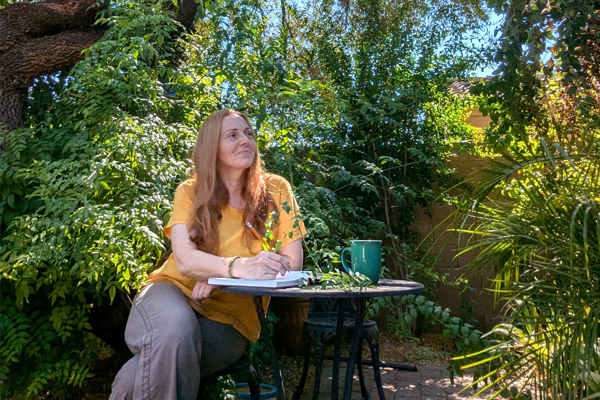 Dee sits in a garden writing in her journal on a garden table.