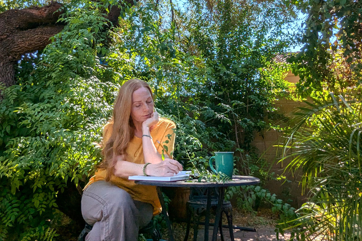 Dee sits in a garden writing in a journal.
