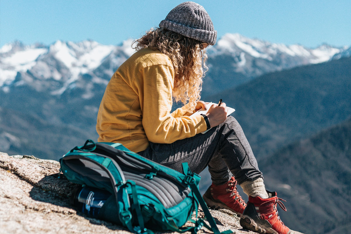 A woman sits on a hill writing in her travel journal.