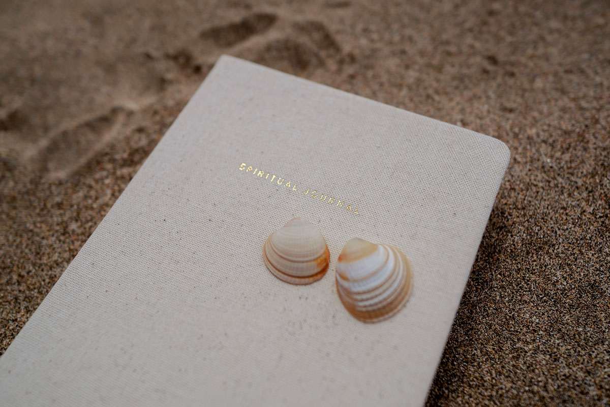 A calendar lays in the sand with two seashells on top.