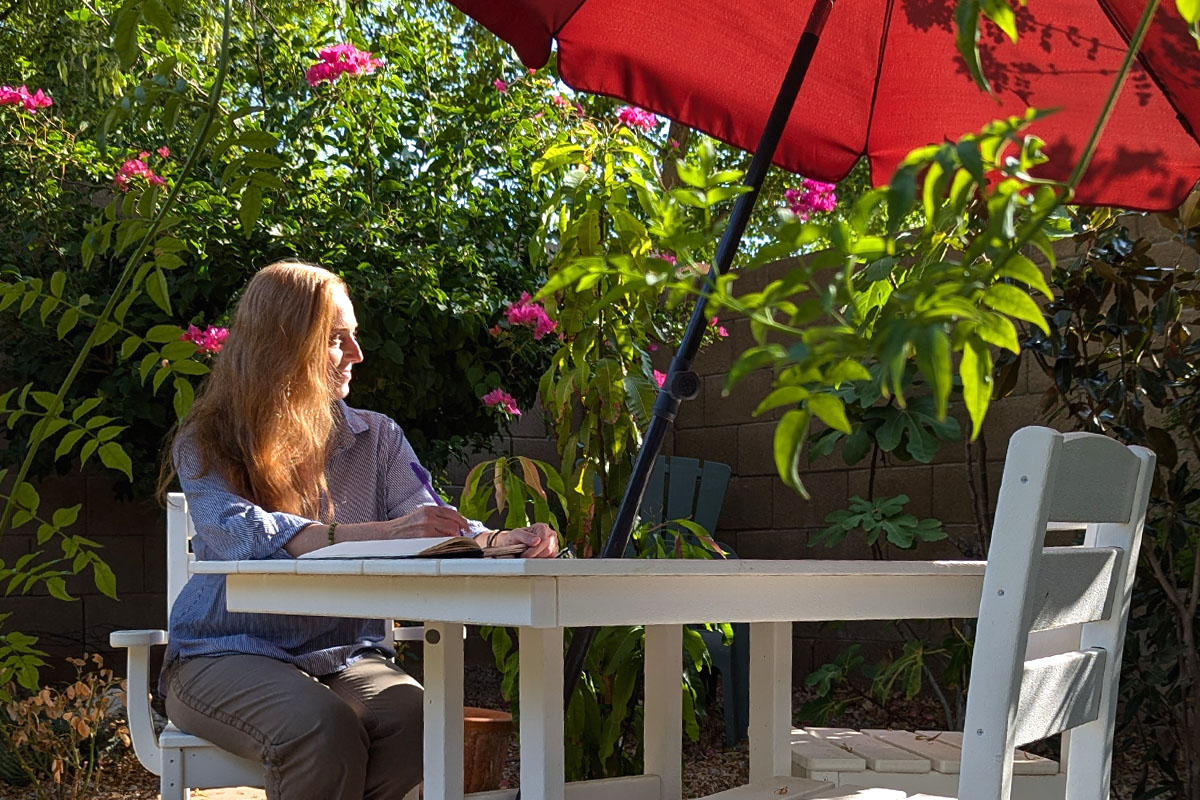Dee sits in the garden with a journal under a red umbrella. 