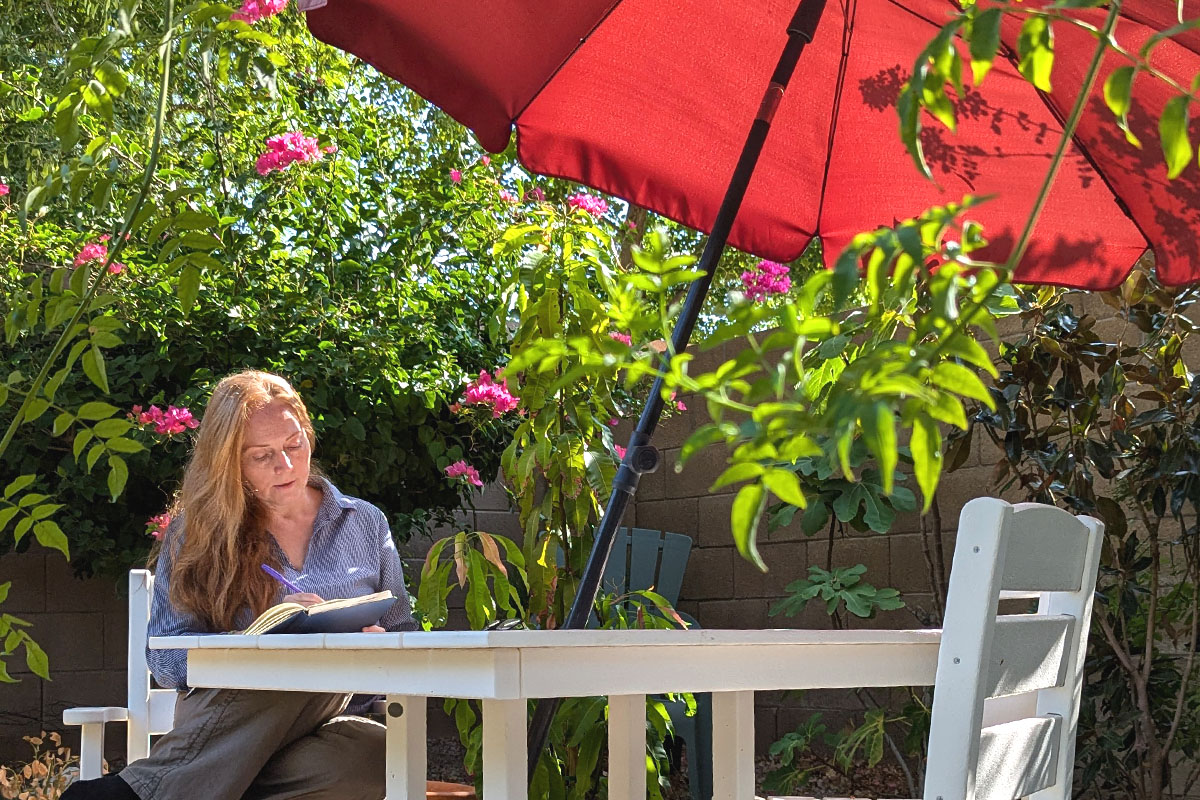 Dee sits in a garden writing in her journal under a red umbrella surrounded by greenery.