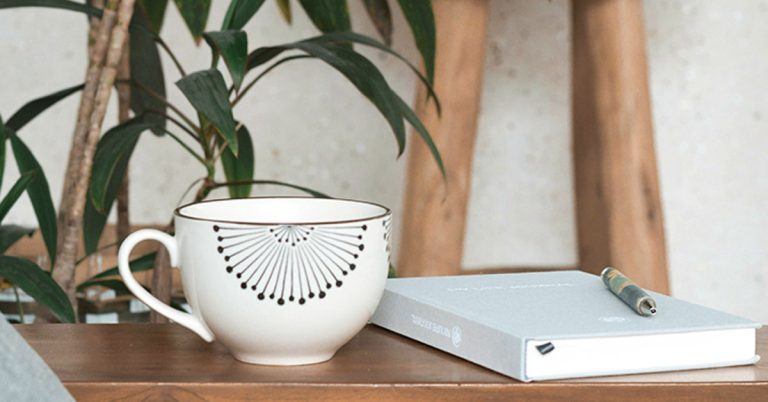 A grey journal lays on a wooden table next to a white coffee cup and green plants in the background.