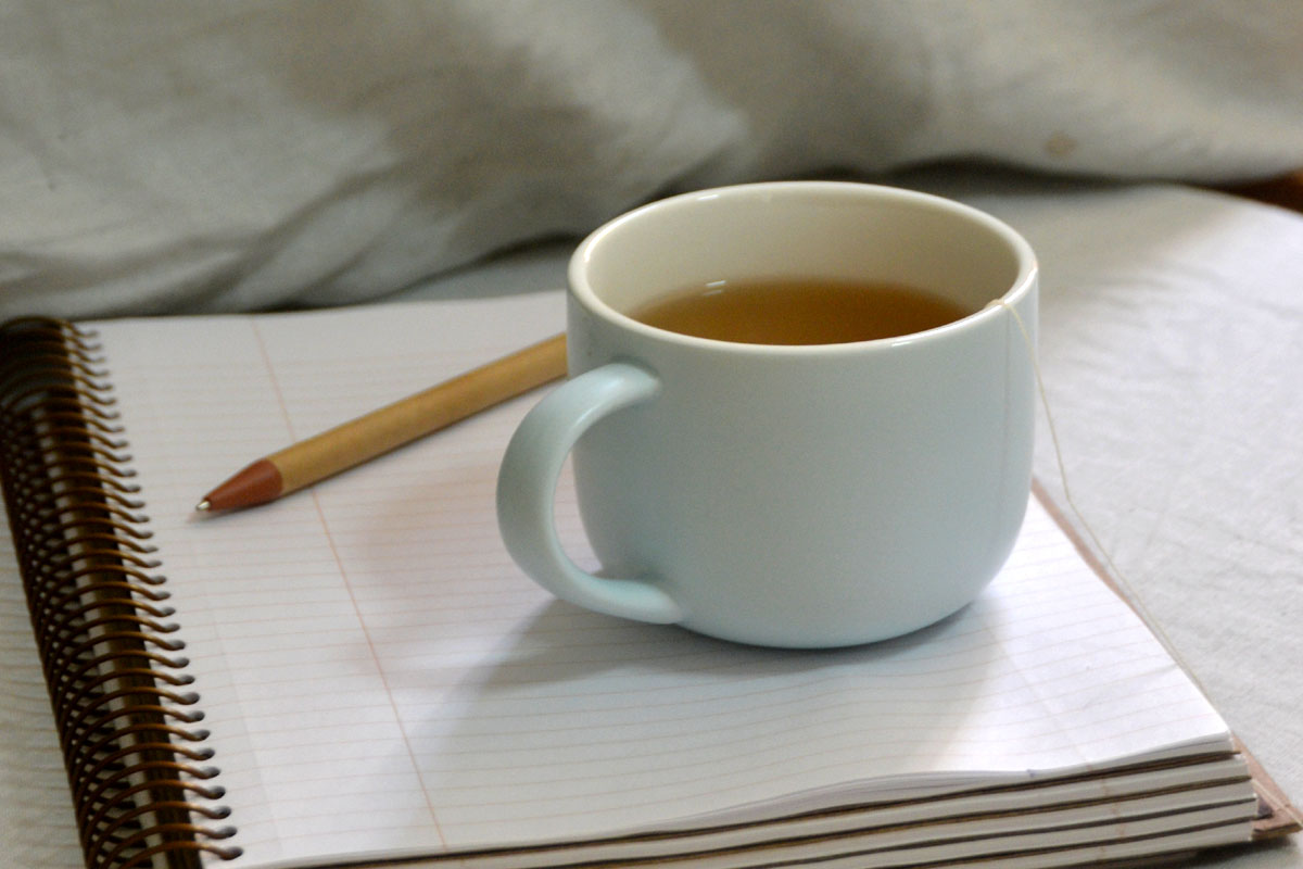 A bullet journal lays on a bed next to a pen and a cup of tea. 