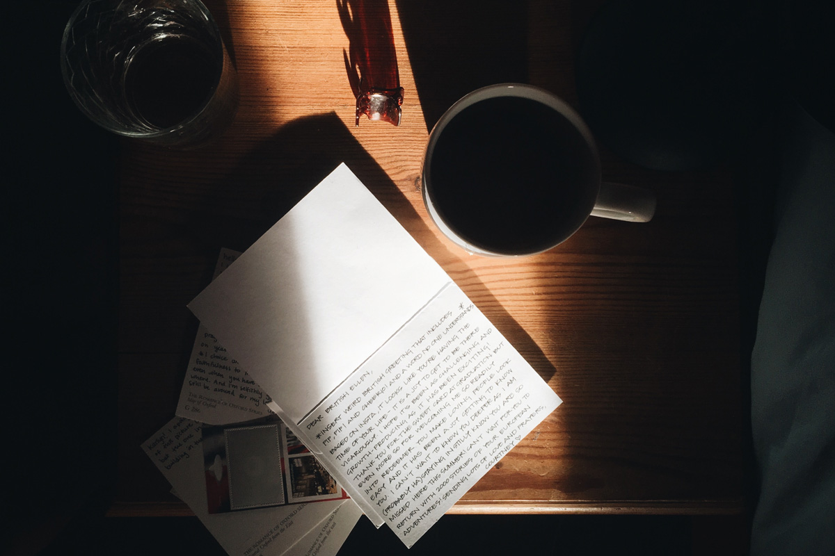 A notebook lays on a wooden table next to a dark cup of coffee. A bright streak of sunshine comes in through the window.