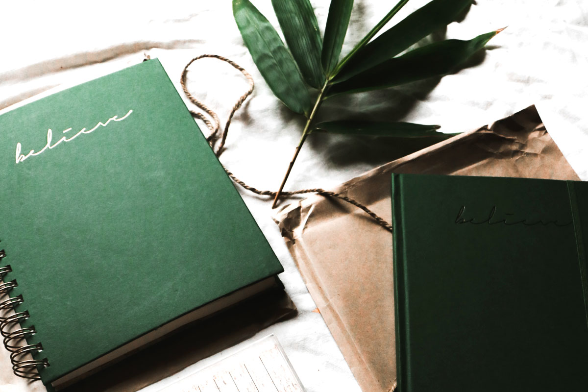 Green notebooks and journals lay on a desk next to green leaves and pens.