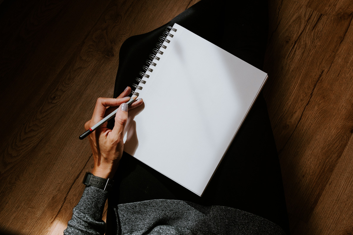 A woman writes in a white notebook against a dark brown background.