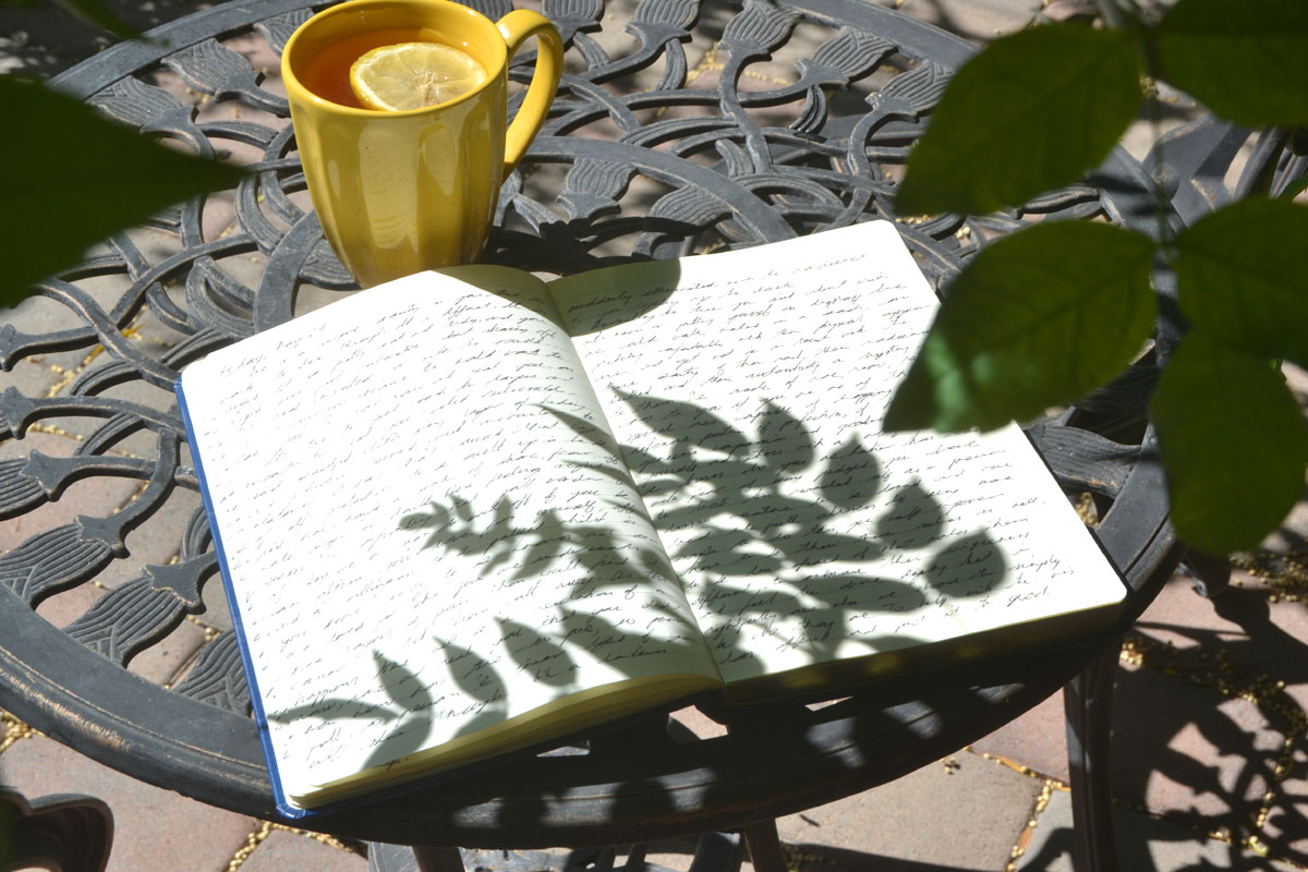 A journal lays open on a garden table with shadows of leaves over its pages. 