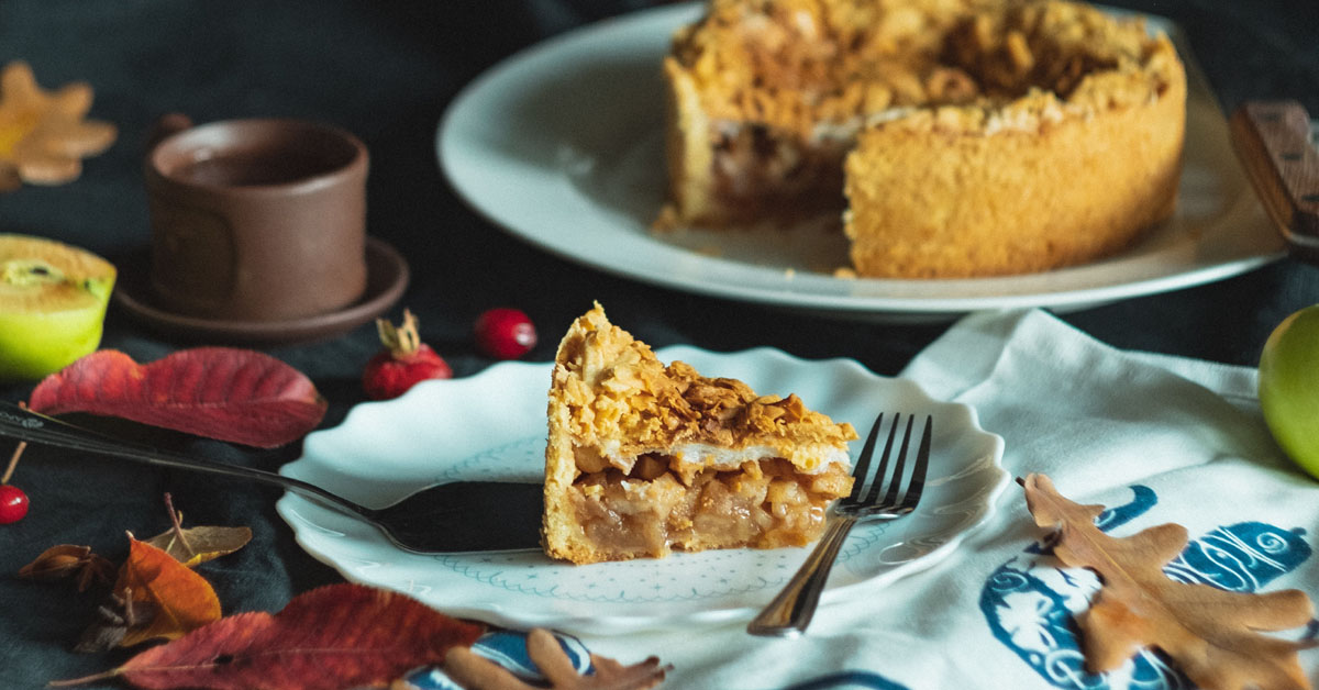 A slice of apple pie lays on a table surrounded by cloths and leaves.