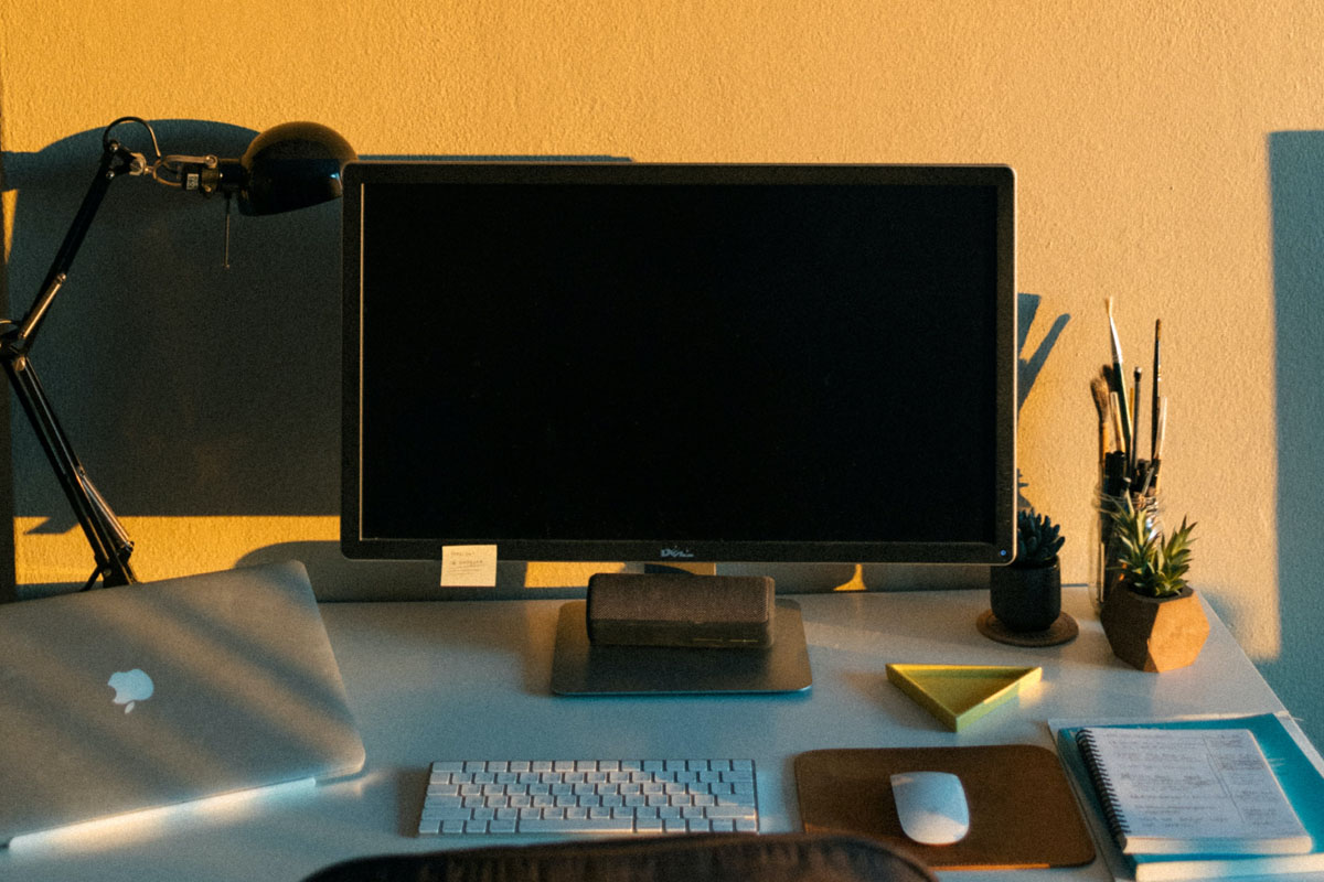 A computer stands on a desk against a wall lit up in sunshine. 