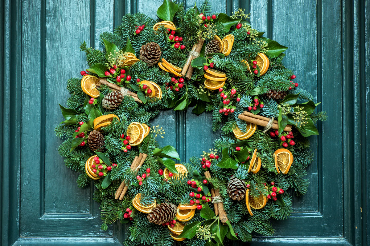 A wreath hangs on a blue door decorated with leaves and oranges. 