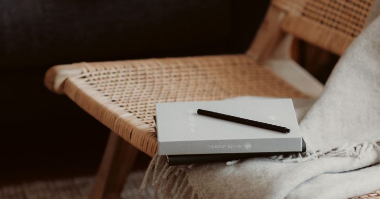 A grey journal lays on a modern wicker chair with a grey throw nearby.