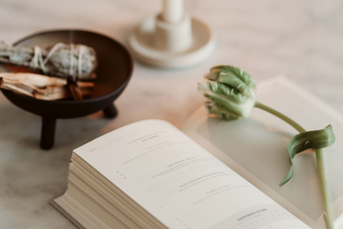 A journal lays open next to burning incense and a green flower. 