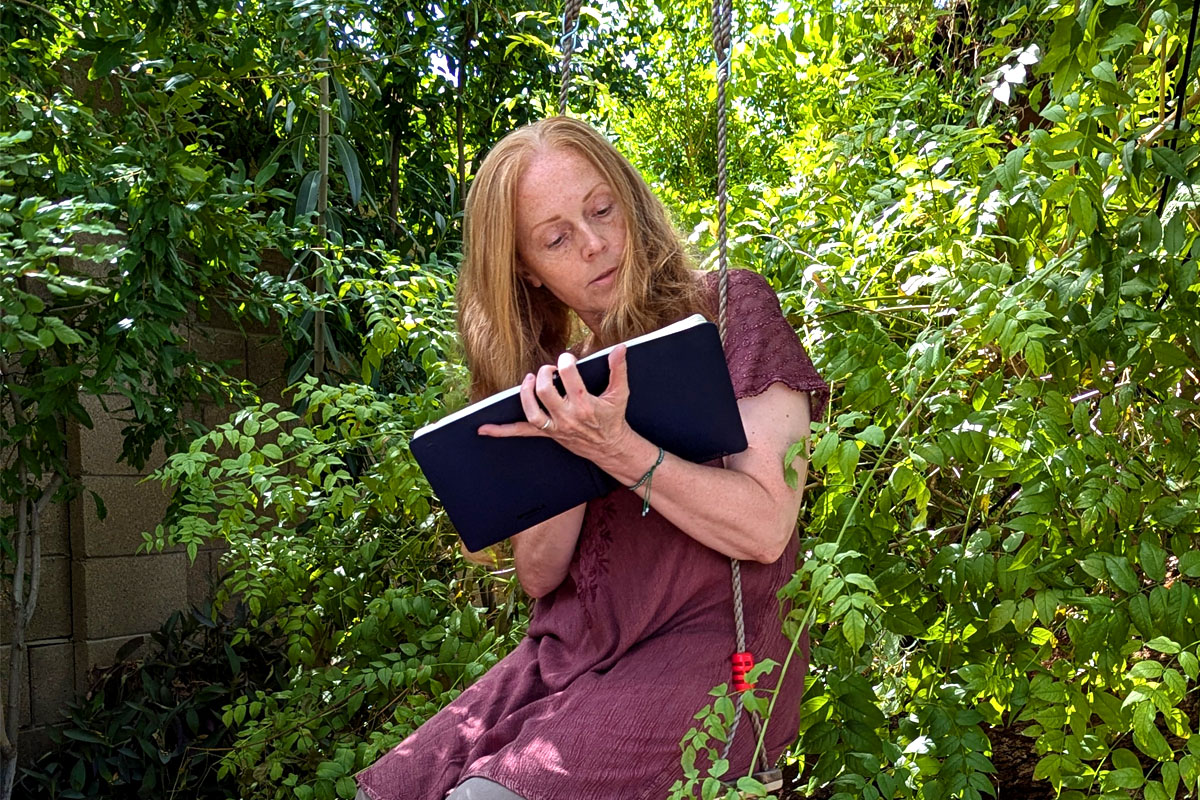 Dee sits on a swing surrounded by greenery and writes in her journal.