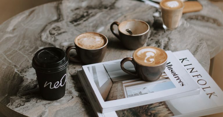 A pile of magazines lay on a table with cups of coffee.