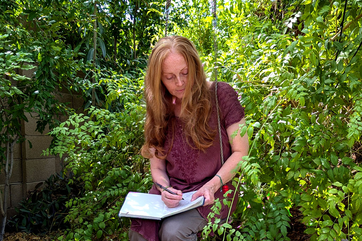 Dee sits on a swing surrounded by greenery writing in her journal.