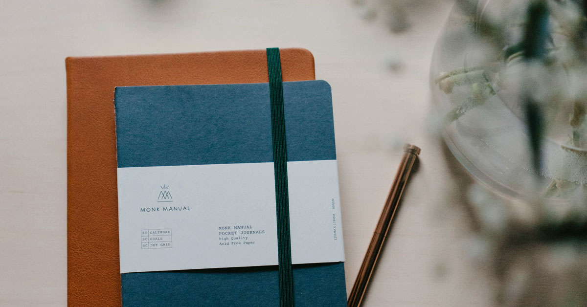Notebooks with daily journal prompts lay on a desk next to a pen and a vase of greenery.