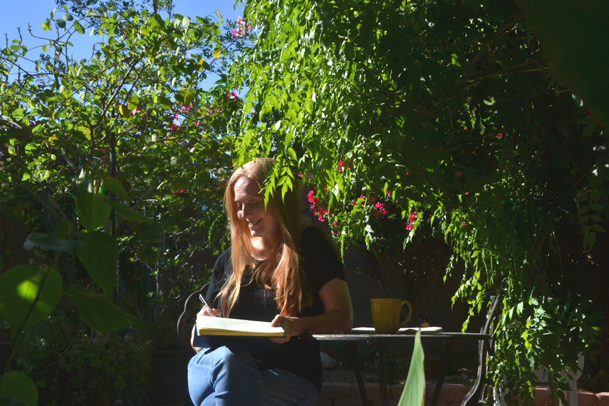 Dee sits in a garden writing in a diary.