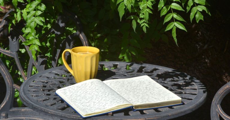 An open notebook lays on a garden table next to some greenery and a cup of tea.