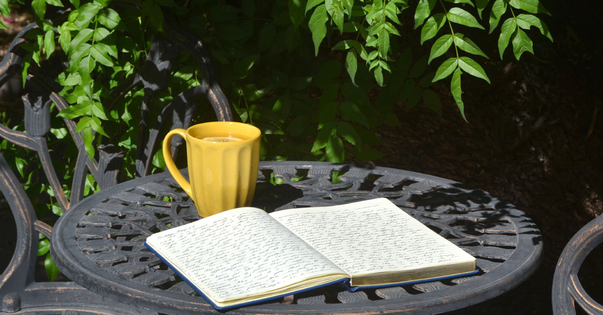 An open notebook lays on a garden table next to some greenery and a cup of tea.