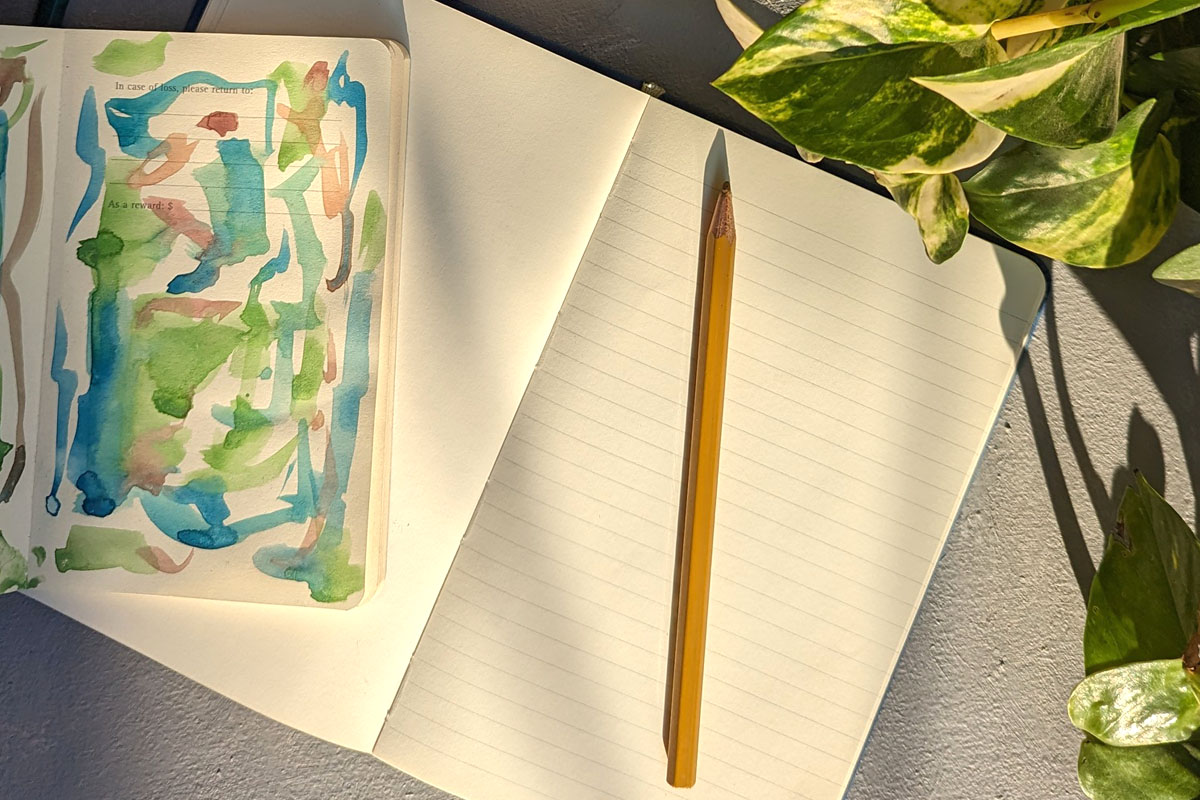 A gratitude journal lays in the sun next to a plant with a pencil on top.