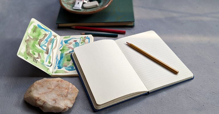 An empty gratitude journal lays open on a grey desk next to a smaller notebook filled with watercolor sketches, a yellow pencil and a large paperweight rock.