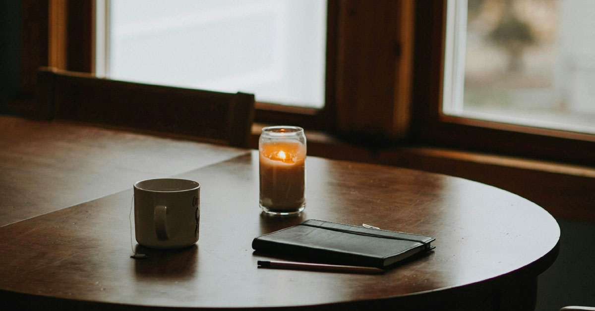 A notebook lays next to a lit candle on a dark wooden desk against a dark room with bright windows.