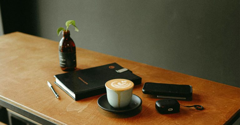 A notebook with April journal prompts lays on a wooden table next to a vase with a twig of greenery and a white coffee mug with latte art.