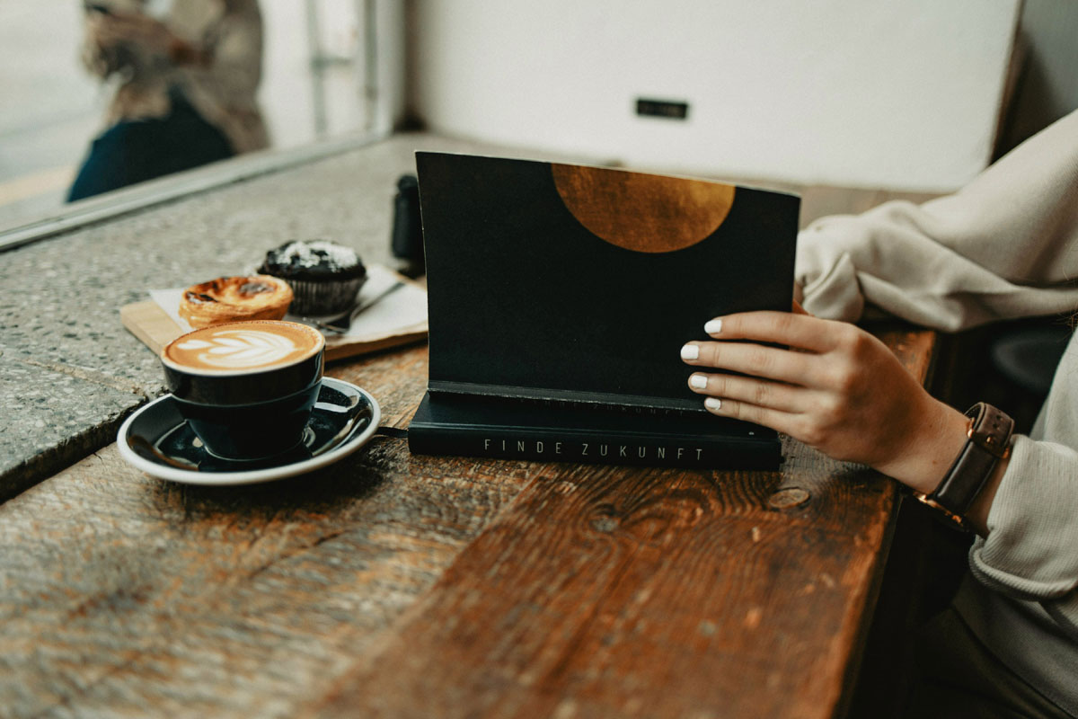A woman sits at a wooden table opening a notebook with a cup of coffee beside her during a digital detox.
