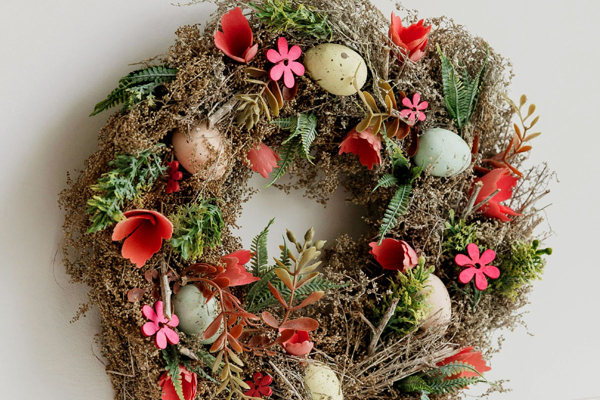 A wreath with eggs and pink and red flowers hangs on a white door.