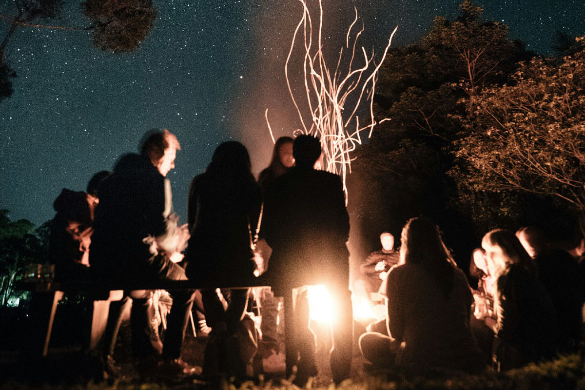 People stand around a bonfire in a forest at night.