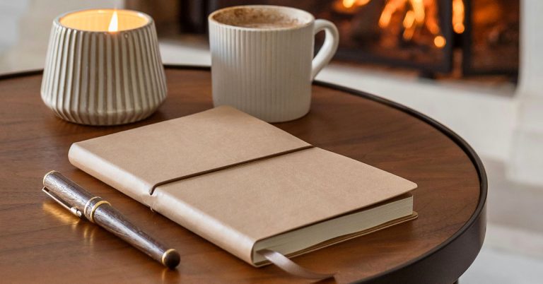 A notebook full of journaling prompts for January lays on a table next to mugs of coffee and a fireplace in the distance.
