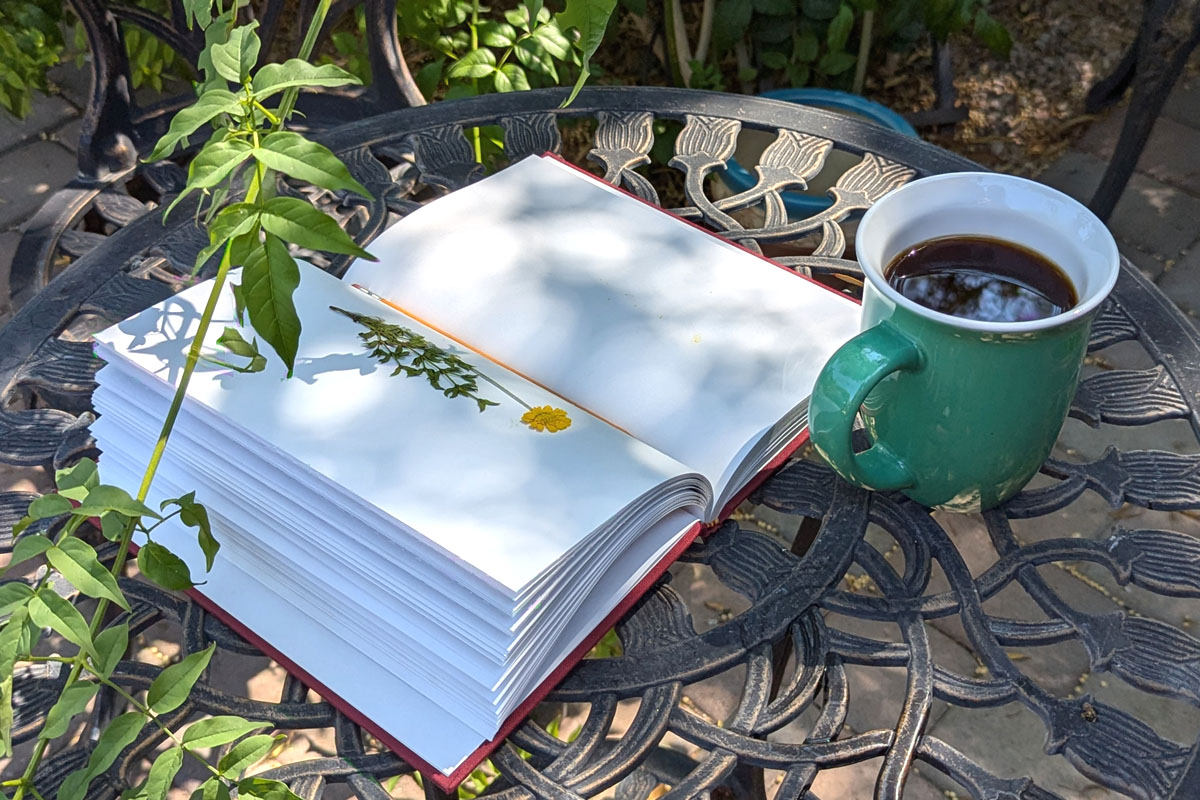 A journal lays on a table in a garden next to a cup of coffee surrounded by greenery.