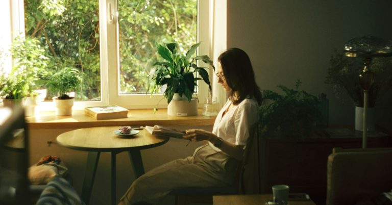 A woman sits next to a window reading a book with a potted plant resting on a table.