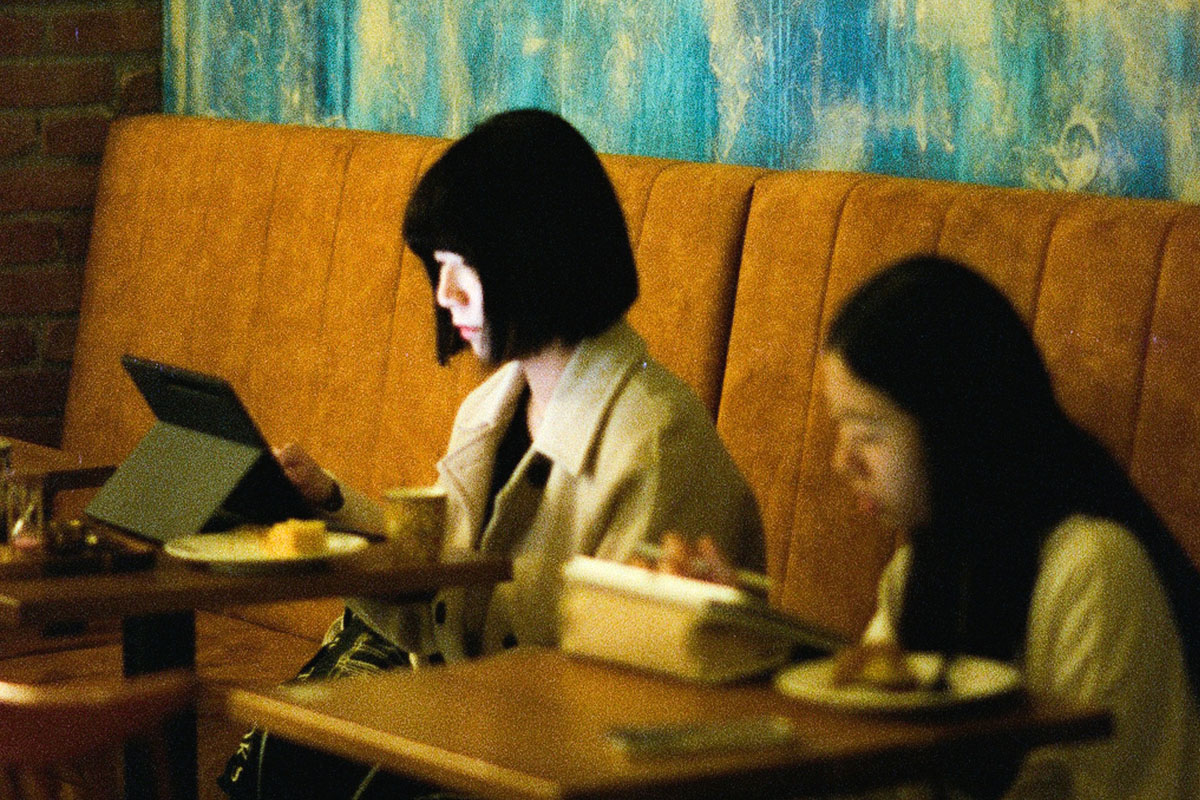 Two women sit at a cafe looking at their laptop and phone screens.