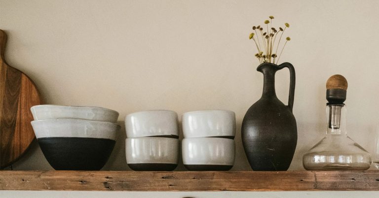 A vase, cups and plates lined up on a wooden kitchen shelf as an example of simple living.