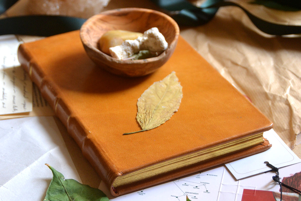 A yellow leather journal lays on a table alongside a wooden bowl and leaves.