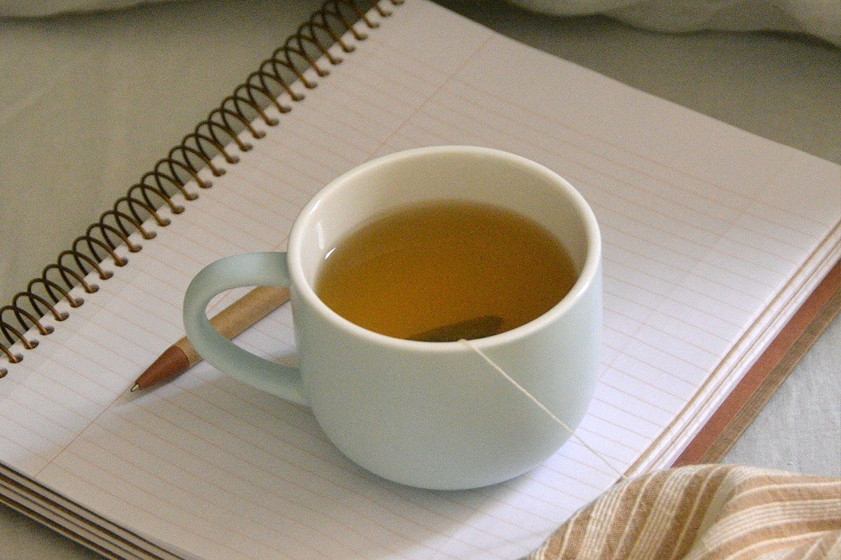 A gratitude journal lays open on a bed with a cup of green tea.