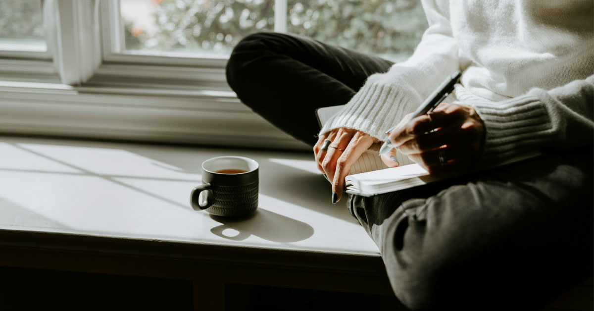 A woman sits on a window sill in the sun with a cup of coffee and a blank journal in the morning.
