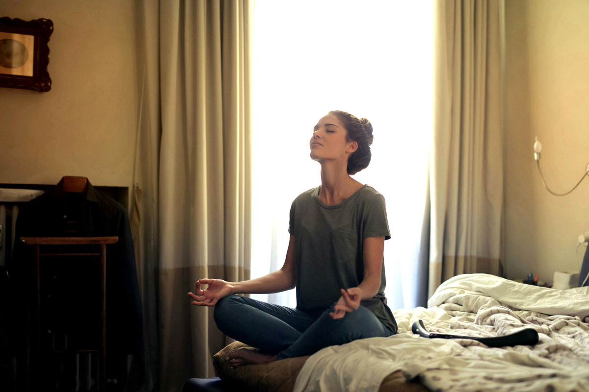 A woman sits on her bed and mediates with curtains and a tall window behind her.