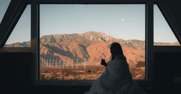 A woman sits at a window with a cup of coffee looking out at a view of some red mountains in California as part of her morning routine.