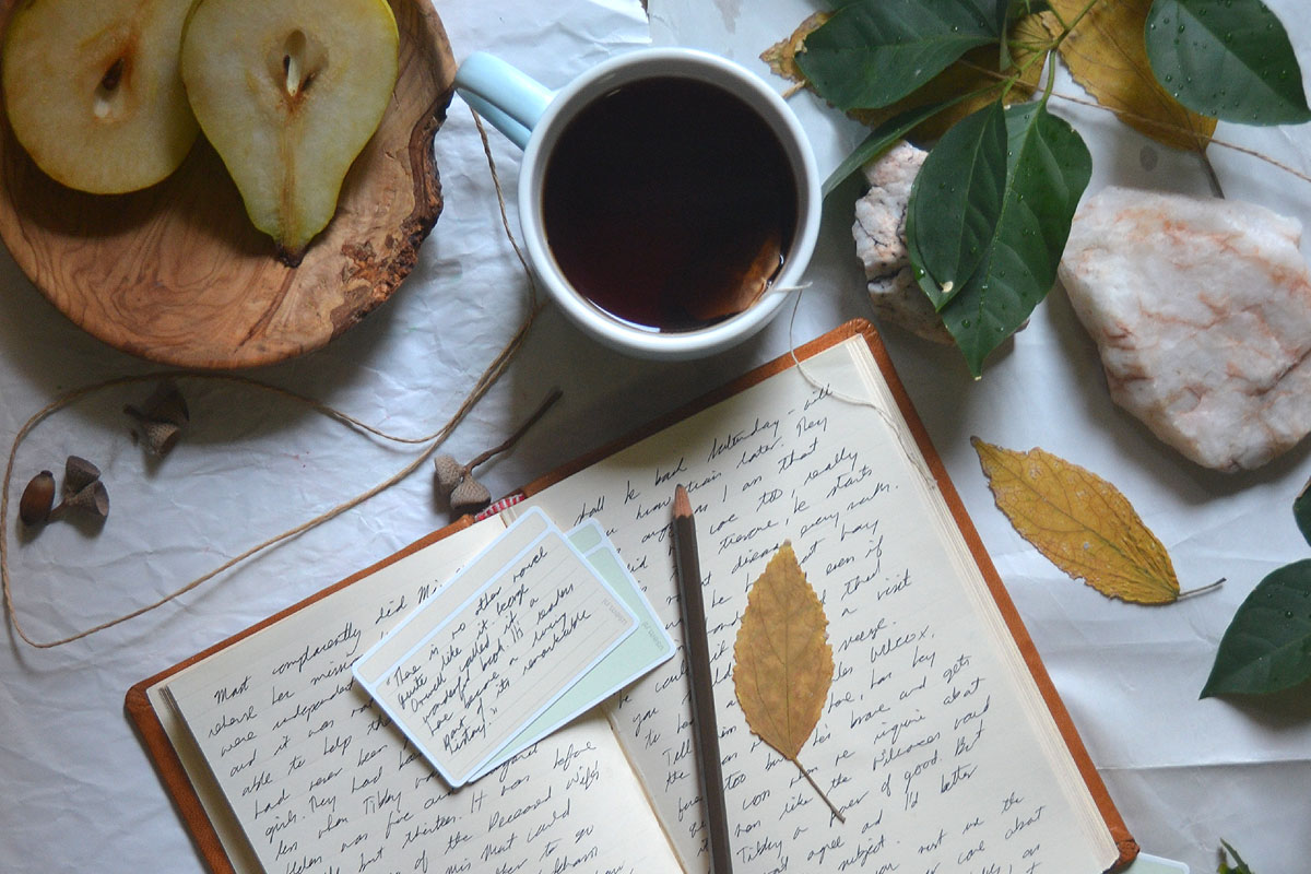 A journal lays on a table with leaves, a cup of tea and a bowl of fruit. 