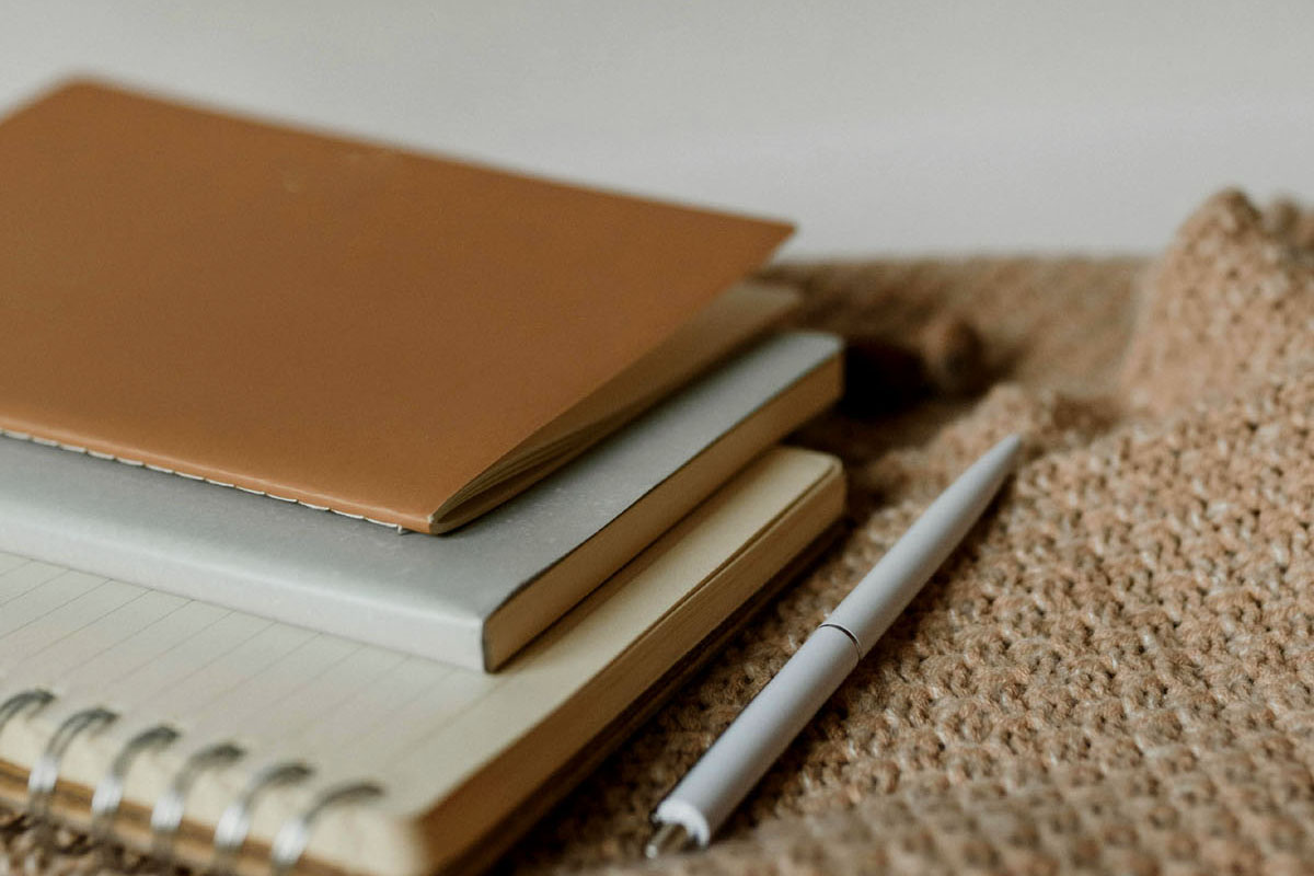 A stack of notebooks lay on a bed alongside a beige blanket. 