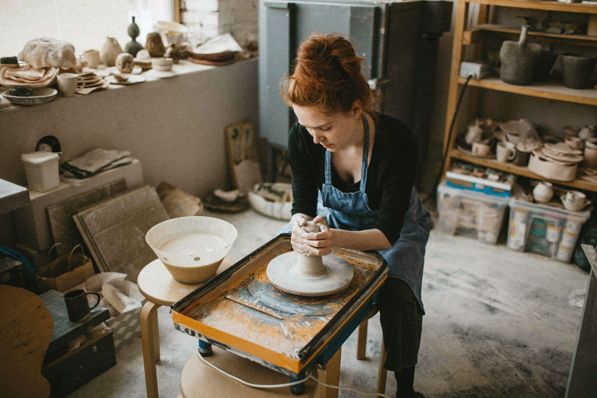A woman sits at a pottery wheel making a tall vase.
